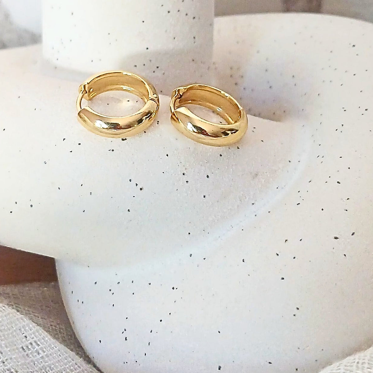 A pair of polished gold huggie hoop earrings displayed on a white speckled jewelry bust.