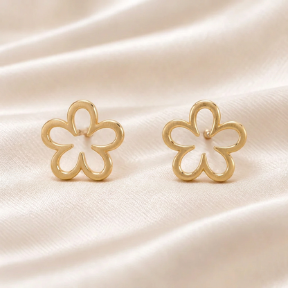 A pair of gold flower-shaped stud earrings with openwork petals, displayed on a soft, draped cream-colored fabric background.