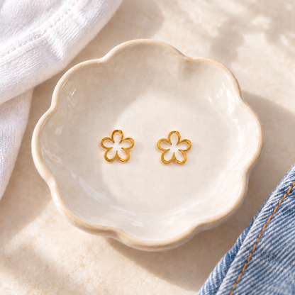 A pair of small gold flower-shaped stud earrings displayed in a scalloped cream ceramic dish, styled with a white knit fabric and blue denim on a light beige surface.