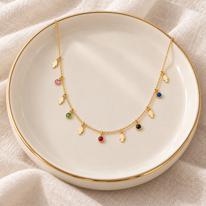A delicate gold necklace with small hand shaped charms and colorful gemstone accents, displayed on a white ceramic plate with a gold rim over a soft beige fabric background.
