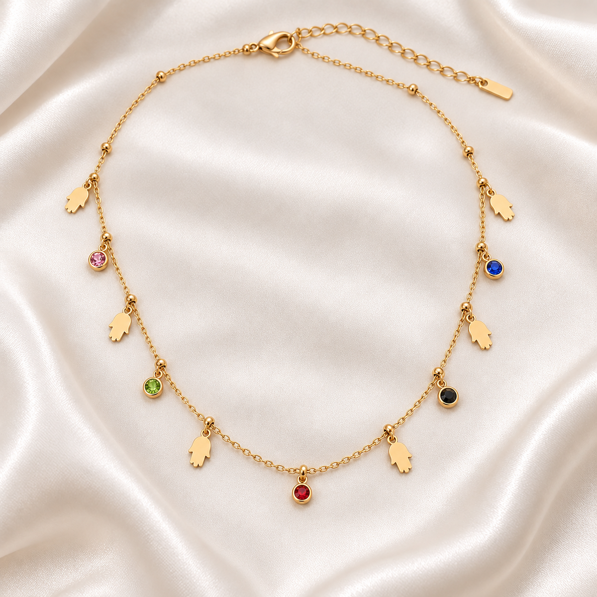A gold-toned necklace with multiple dangling charms, including five hand-shaped charms and several small oval gemstone charms in pink, blue, green, black, red, and white, displayed on a white satin fabric background.