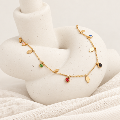 A delicate gold chain necklace with small hand charms and several round gemstone charms in green, red, blue, and black, displayed on a white speckled vase
