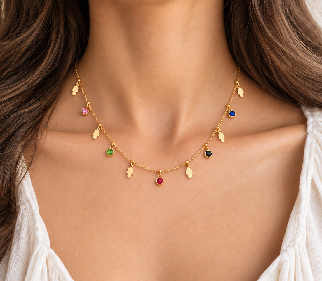 A delicate gold necklace with hand of hamsa charms and colorful round gemstone accents, worn on a model's neck over a white top.