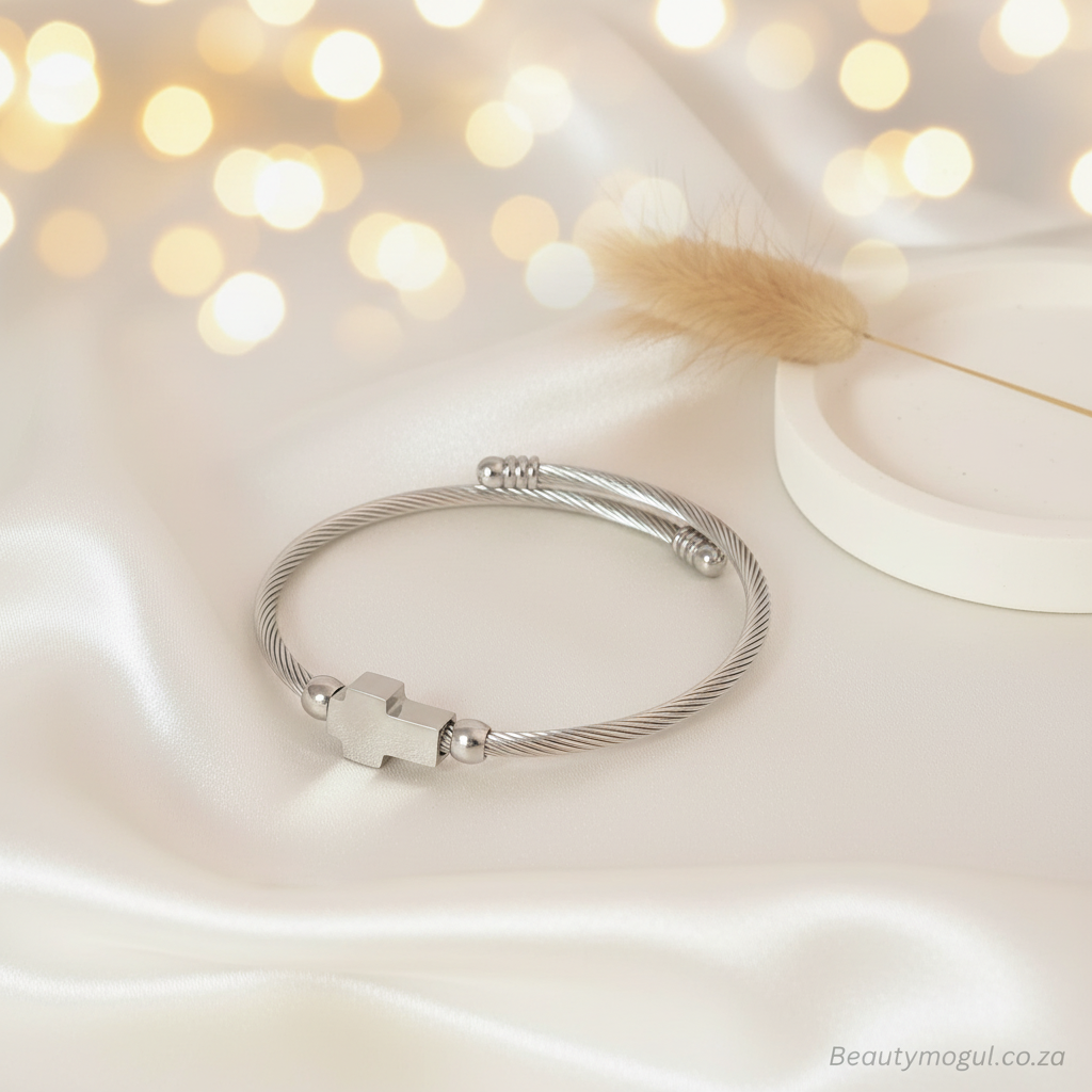 A silver-toned cable-wire bangle bracelet with a small cross charm and bead accents, displayed on draped satin fabric with warm bokeh lights and a decorative dried flower in the background.