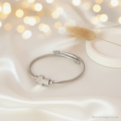 A silver-toned cable-wire bangle bracelet with a small cross charm and bead accents, displayed on draped satin fabric with warm bokeh lights and a decorative dried flower in the background.