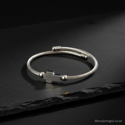A silver-toned twisted cable bangle bracelet with a central cross-shaped charm and small bead accents, displayed on a dark textured slate surface.