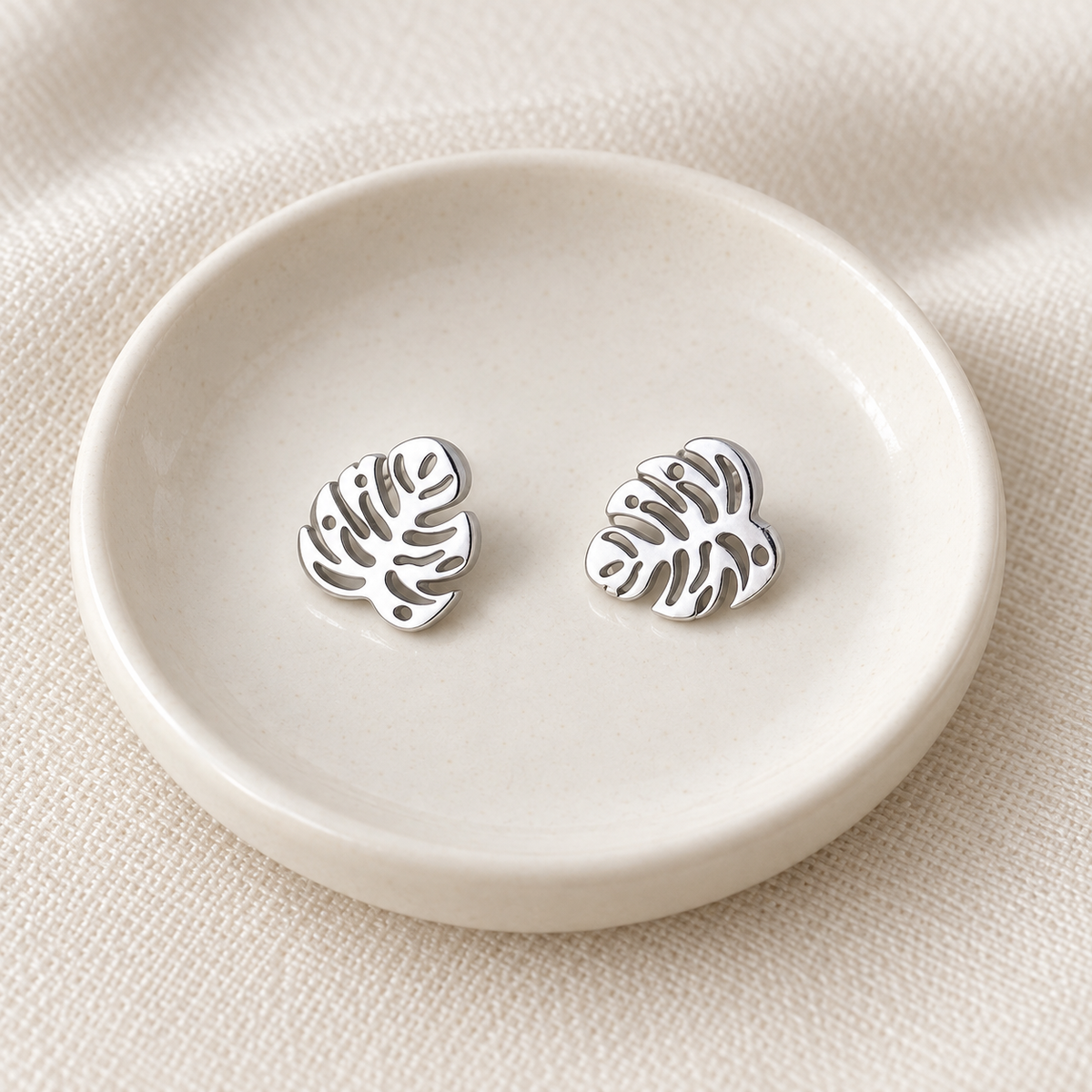 A pair of silver-toned leaf-shaped stud earrings displayed on a small round off-white dish over a textured beige fabric background.