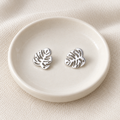A pair of silver-toned leaf-shaped stud earrings displayed on a small round off-white dish over a textured beige fabric background.
