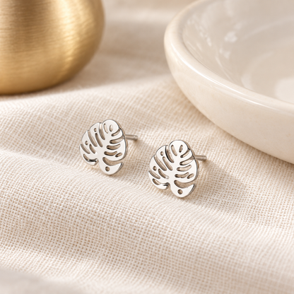 Silver monstera leaf stud earrings styled with cream fabric and minimal boutique accessories.