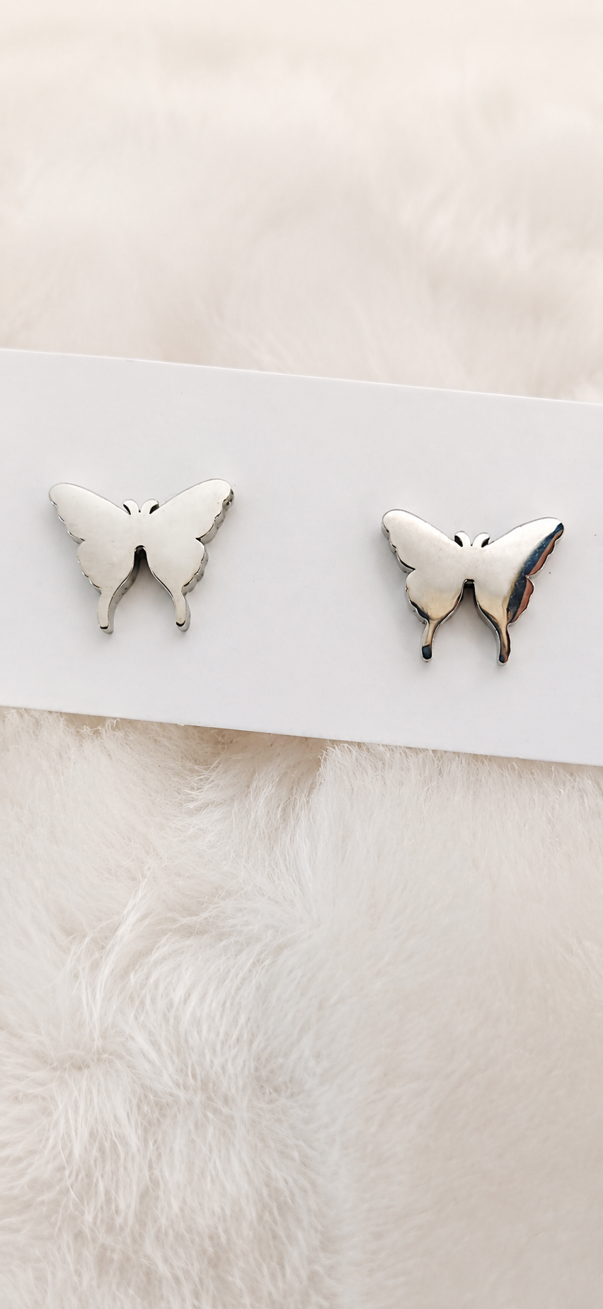 A pair of white butterfly stud earrings with silver-toned edges, displayed on a white card over a soft white faux fur background.