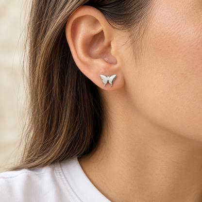 A close-up of a woman's ear wearing a small silver butterfly stud earring, with her hair and shoulder visible against a soft light background.