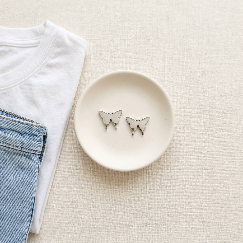 A pair of small butterfly-shaped stud earrings in silver-tone metal with white enamel details, displayed on a small white dish next to folded white and blue clothing on a light beige textured background.
