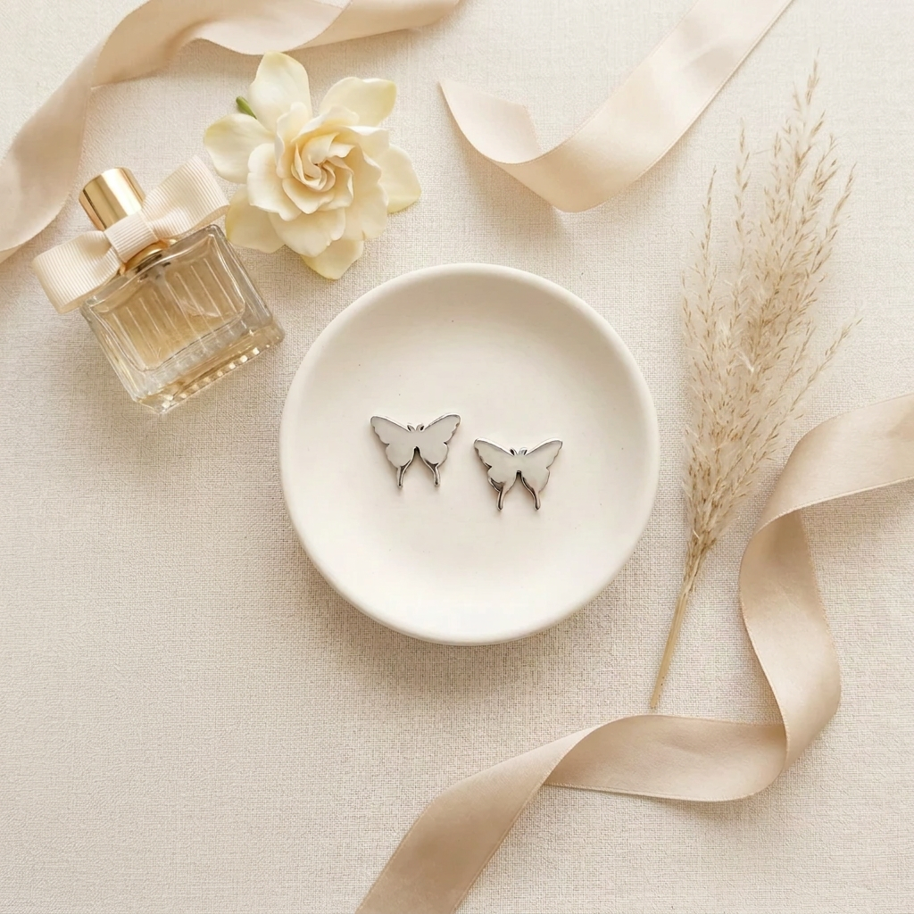 A pair of silver butterfly stud earrings displayed on a small white dish, styled in a flat-lay with a perfume bottle, a cream flower, dried pampas grass, and beige ribbons on a textured cream fabric background.