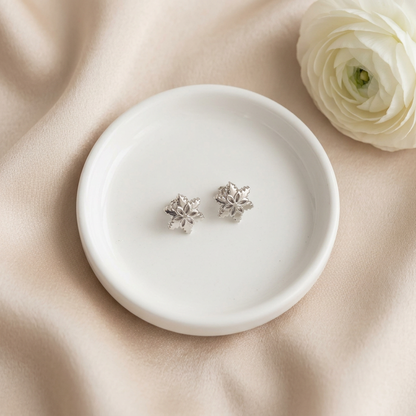 Silver-tone snowflake stud earrings on an ivory jewellery dish with champagne satin and a white flower