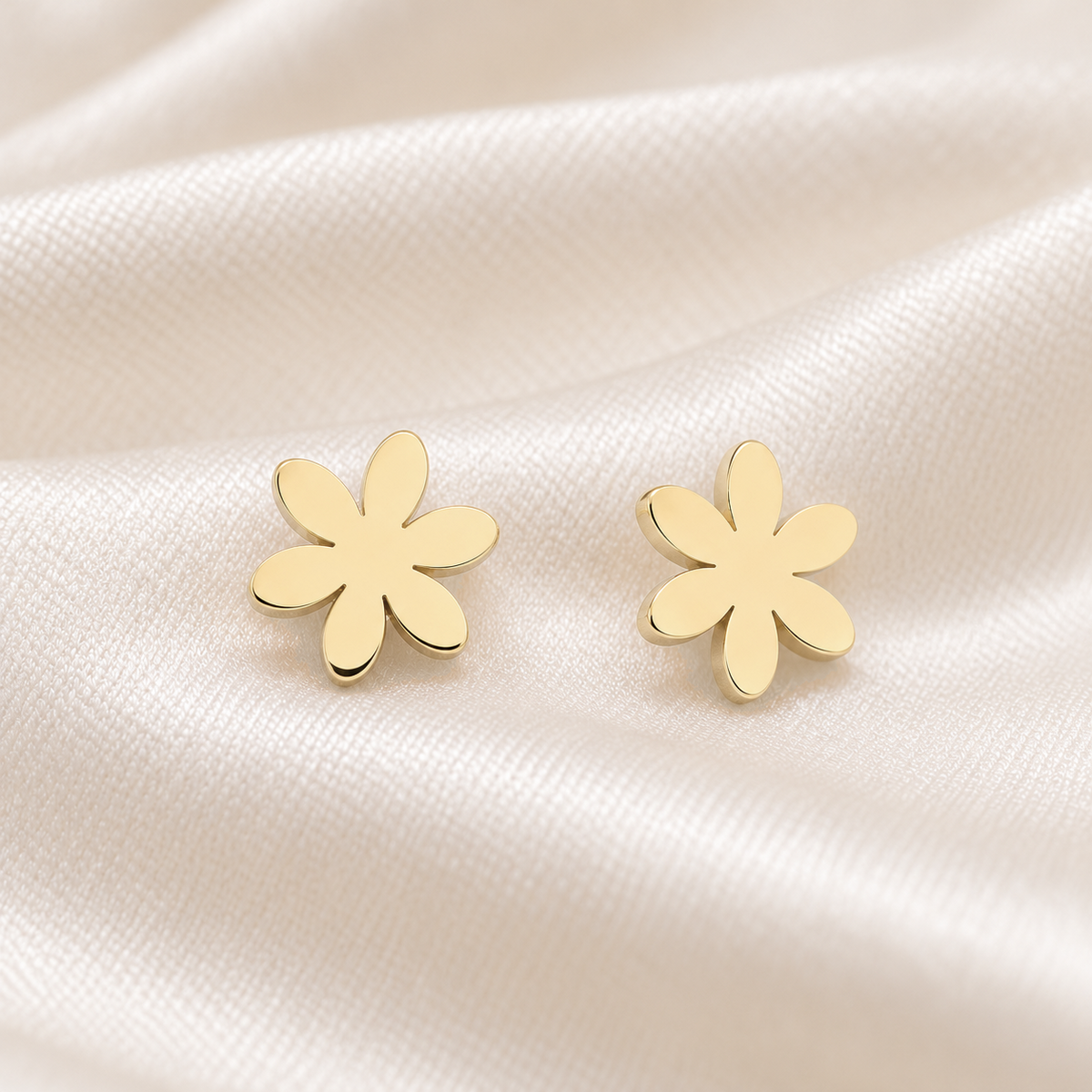 A pair of small gold flower-shaped stud earrings resting on soft cream-colored satin fabric.