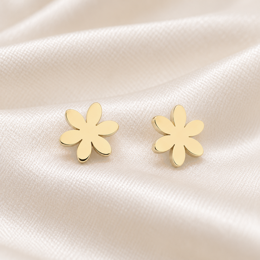 A pair of small gold flower-shaped stud earrings resting on soft cream-colored satin fabric.