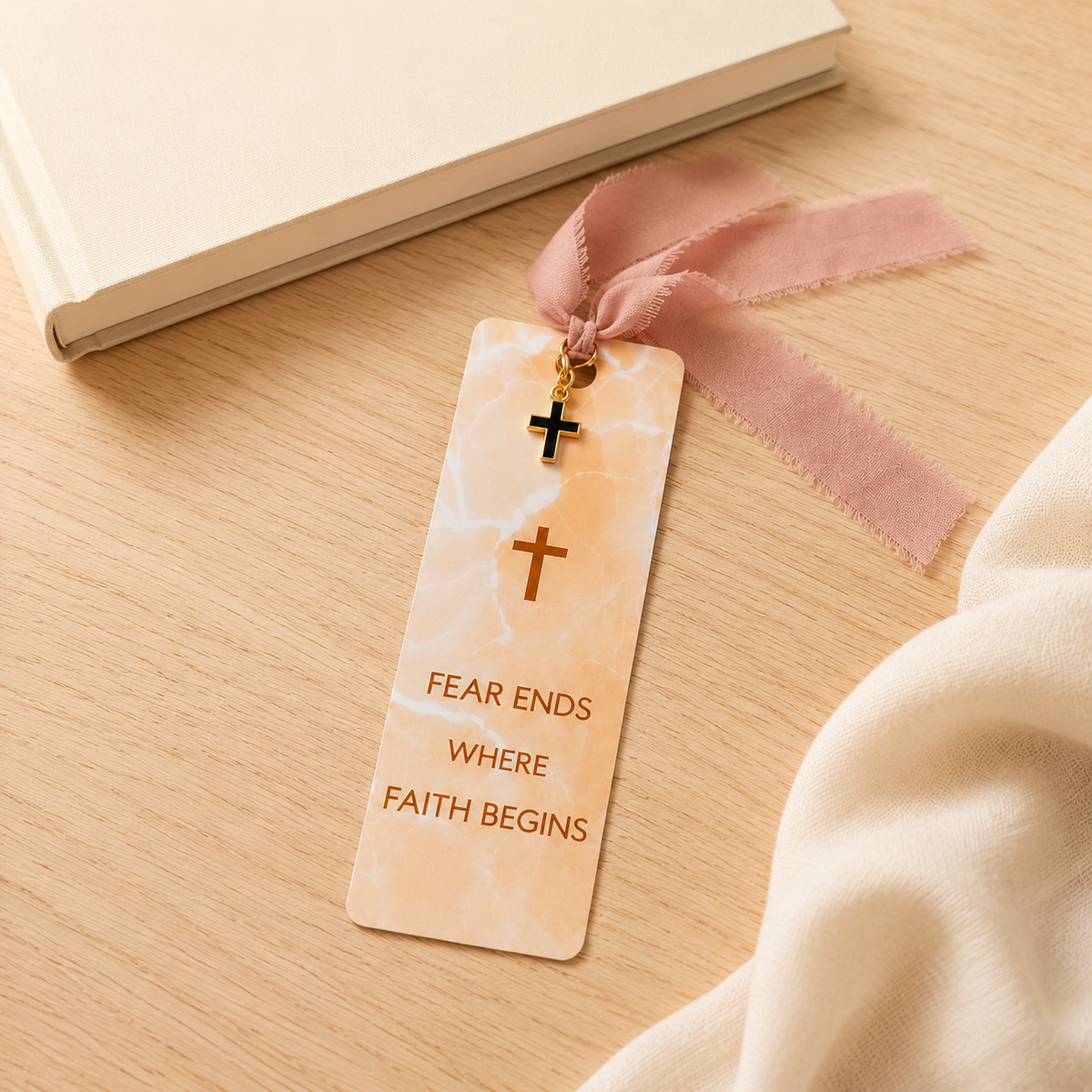 A cream-colored marble bookmark with a pink ribbon and a small gold cross charm, lying on a light wood desk next to a book and a soft cream fabric. The bookmark features the text 'FEAR ENDS WHERE FAITH BEGINS'.