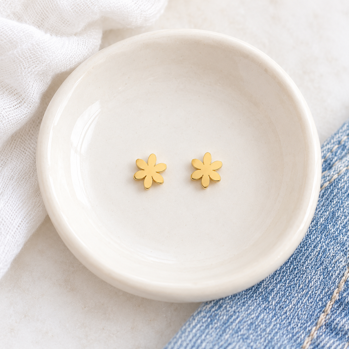 A pair of small yellow gold flower-shaped stud earrings resting in a shallow white ceramic dish, with soft white fabric and blue denim visible around the dish.