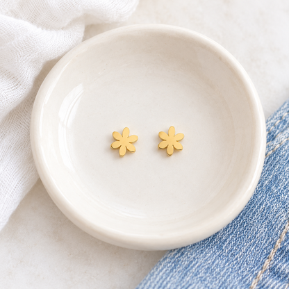 A pair of small yellow gold flower-shaped stud earrings resting in a shallow white ceramic dish, with soft white fabric and blue denim visible around the dish.