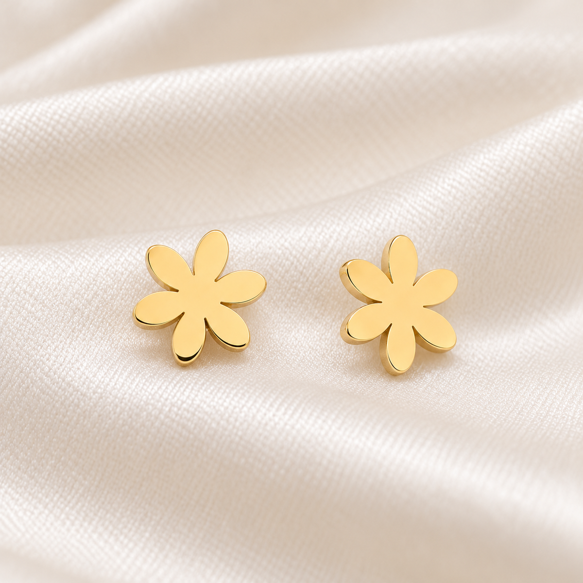 A pair of gold flower-shaped stud earrings resting on a soft, draped white satin fabric background.