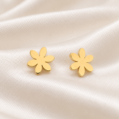 A pair of gold flower-shaped stud earrings resting on a soft, draped white satin fabric background.