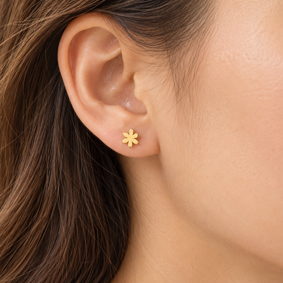 A close-up of a woman's ear wearing a small gold flower stud earring, with her brown hair and skin visible against a soft light background.