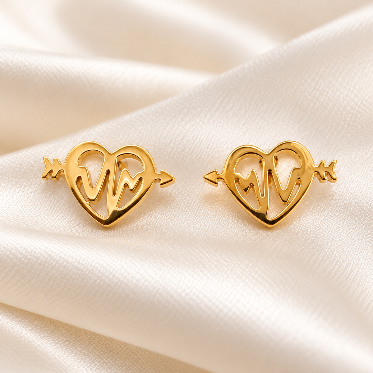 A pair of gold heart-shaped stud earrings with arrow ends and an EKG line design in the center, displayed on a cream satin fabric background.