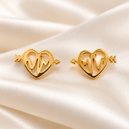 A pair of gold heart-shaped stud earrings with arrow ends and an EKG line design in the center, displayed on a cream satin fabric background.