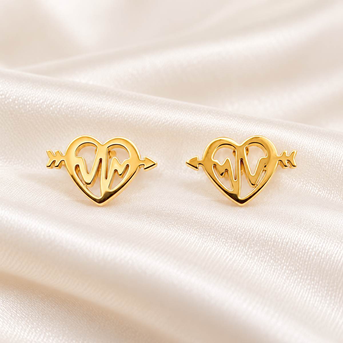 A pair of gold heart-shaped stud earrings with an openwork design, displayed on a draped cream satin fabric background.