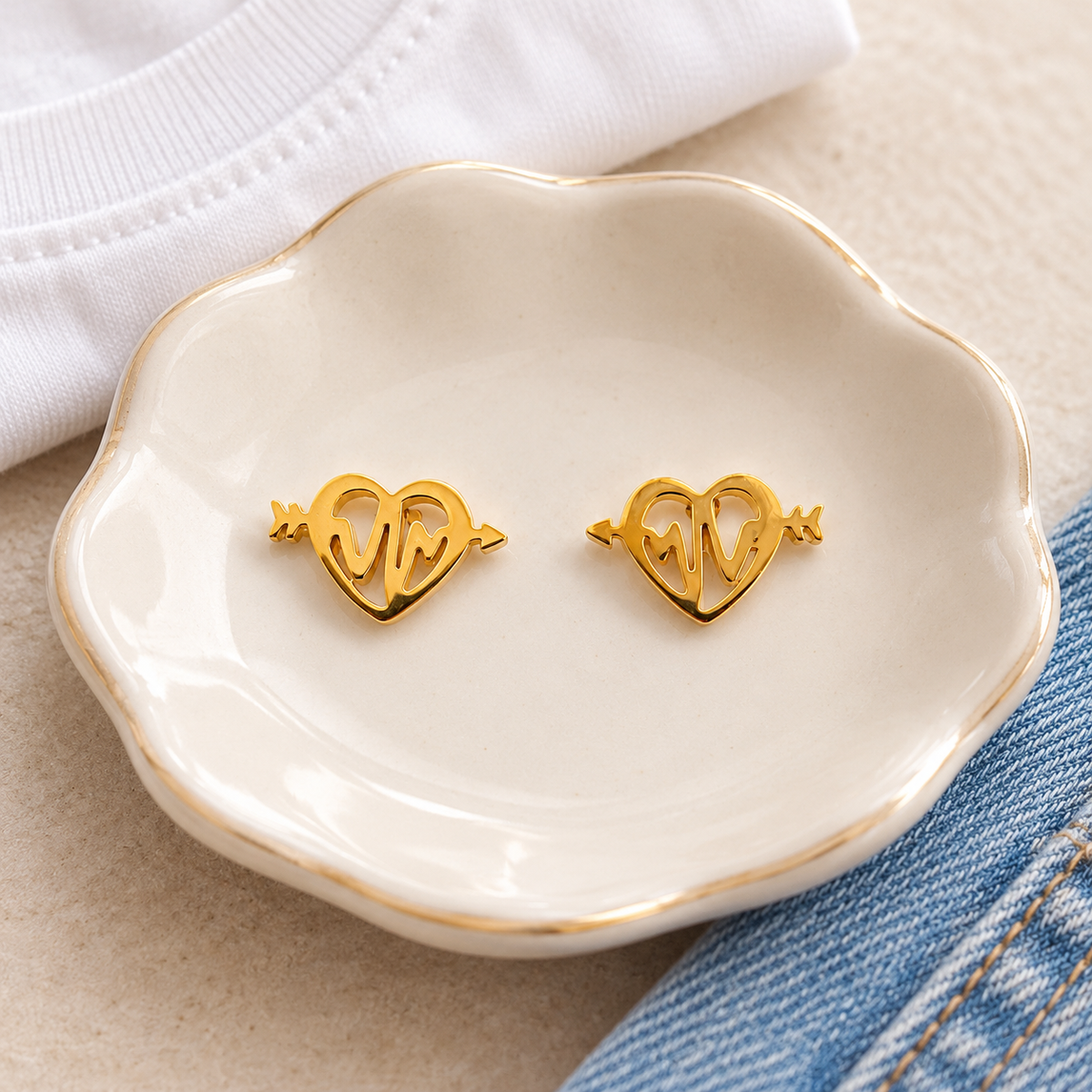 A pair of gold heart-shaped stud earrings with arrow details, displayed on a small white ceramic dish with gold trim, styled with a white t-shirt and blue denim.