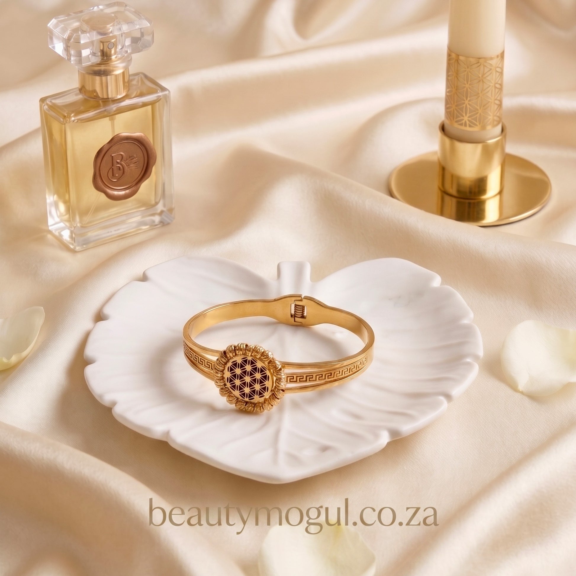 Gold bracelet on a white shell with a perfume bottle and candle on a beige fabric background, featuring 'beautymogul.co.za'.