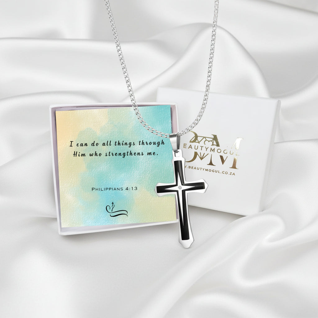 A silver cross pendant necklace with black enamel detailing, displayed on a silver chain. It is presented on a white satin background alongside a decorative card with a Bible verse and a branded gift box from Beauty Mogul.