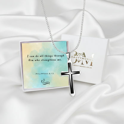 A silver cross pendant necklace with black enamel detailing, displayed on a silver chain. It is presented on a white satin background alongside a decorative card with a Bible verse and a branded gift box from Beauty Mogul.