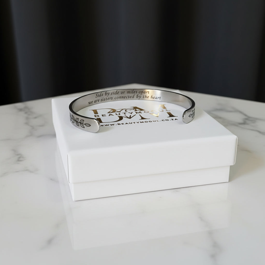 A silver engraved cuff bracelet with the words 'Side by side we make sense, we are sisters connected by the heart'  resting on top of its white presentation box, set on a white marble surface with a dark background.