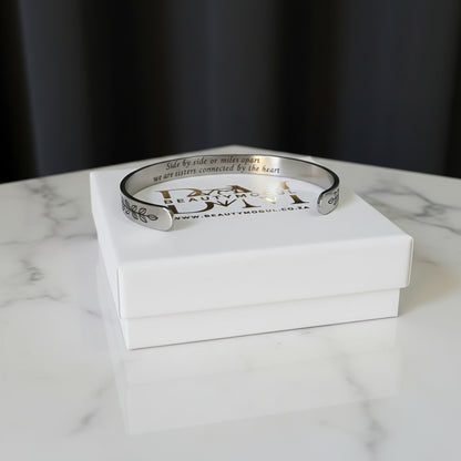 A silver engraved cuff bracelet with the words 'Side by side we make sense, we are sisters connected by the heart'  resting on top of its white presentation box, set on a white marble surface with a dark background.