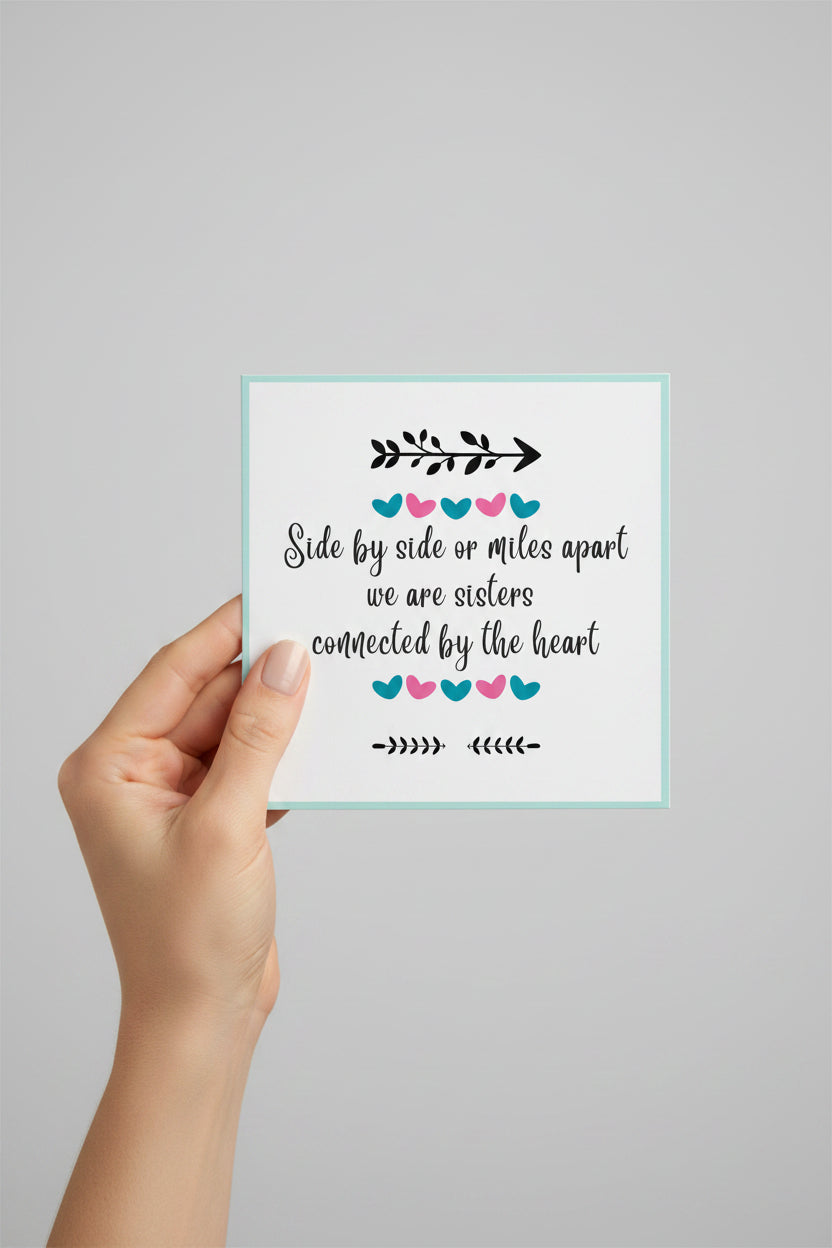 A hand holding a square white greeting card with a light blue border. The card features a black decorative arrow and the text 'Side by side or miles apart we are sisters connected by the heart' surrounded by small pink and blue hearts, against a solid light gray background.