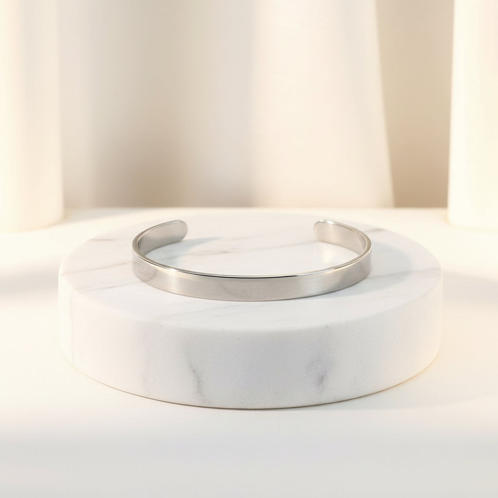A polished silver cuff bracelet displayed on a round white marble pedestal against a soft, warm off-white background.