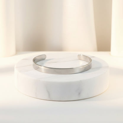 A polished silver cuff bracelet displayed on a round white marble pedestal against a soft, warm off-white background.